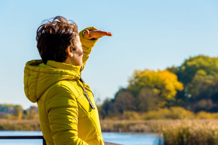A woman in a yellow jacket while traveling looks in the distance, shielding the sun with her hand