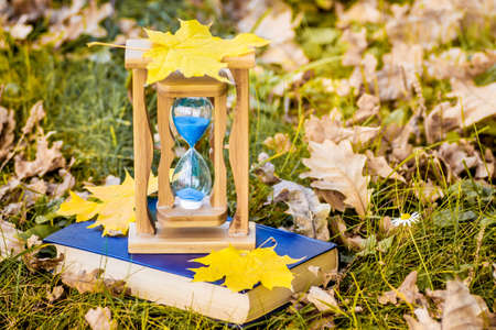 Autumn maple leaves lie on a sand clock and a book in the woodsの写真素材