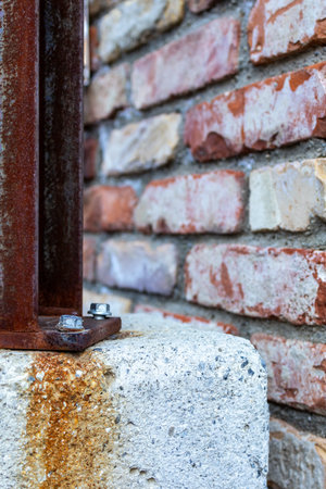 A close-up shot showing a weathered, rusty metal bracket firmly secured to a textured concrete block. The bracket's aged appearance contrasts with the solid, gray concrete. Behind the bracket and concrete, a rustic brick wall adds depth and texture to the scene. The image captures a sense of industrial decay and enduring strength, suitable for themes of construction, repair, or urban landscapes. Shot during a photowalk in Obertauern, Salzburg, Austria in December 2025.の写真素材