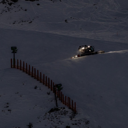 A snowcat diligently grooms a ski slope under the cover of night in the Austrian Alps. The scene is illuminated by the snowcat's headlights, highlighting the freshly groomed snow. A ski lift tower stands in the background, partially obscured by the darkness. The image captures the dedication and effort required to maintain pristine ski conditions, showcasing the beauty and tranquility of a winter landscape.の写真素材