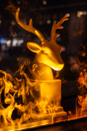 A captivating close-up shot of a reindeer statue illuminated with a warm glow, set against a backdrop of swirling smoke and flickering flames. The statue, with its intricate antlers, stands prominently, creating a striking contrast between the artificial and the natural elements. The warm lighting and dynamic background evoke a sense of festive cheer and mystery. Perfect for holiday-themed projects, advertising campaigns, or artistic compositions.の写真素材