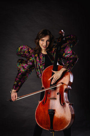 A captivating portrait of cellist Anna Skladannaya against a dark background. She is elegantly dressed in a floral patterned jacket and black pants, holding a cello and bow with grace and focus. The lighting highlights her features and the rich tones of the instrument, creating a dramatic and artistic composition. Perfect for representing music, art, culture, and talent.の写真素材