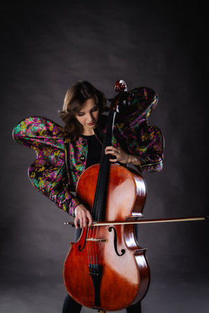 A captivating portrait of cellist Anna Skladannaya against a dark background. She is elegantly dressed in a floral patterned jacket and black pants, holding a cello and bow with grace and focus. The lighting highlights her features and the rich tones of the instrument, creating a dramatic and artistic composition. Perfect for representing music, art, culture, and talent.の写真素材