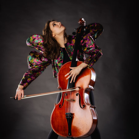 A captivating portrait of a cellist against a dark background. She is elegantly dressed in a floral patterned jacket and black pants, holding a cello and bow with grace and focus. The lighting highlights her features and the rich tones of the instrument, creating a dramatic and artistic composition. Perfect for representing music, art, culture, and talent.の写真素材