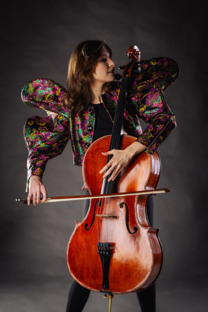 A captivating portrait of cellist Anna Skladannaya against a dark background. She is elegantly dressed in a floral patterned jacket and black pants, holding a cello and bow with grace and focus. The lighting highlights her features and the rich tones of the instrument, creating a dramatic and artistic composition. Perfect for representing music, art, culture, and talent.の写真素材