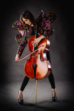 A captivating portrait of cellist Anna Skladannaya against a dark background. She is elegantly dressed in a floral patterned jacket and black pants, holding a cello and bow with grace and focus. The lighting highlights her features and the rich tones of the instrument, creating a dramatic and artistic composition. Perfect for representing music, art, culture, and talent.の写真素材