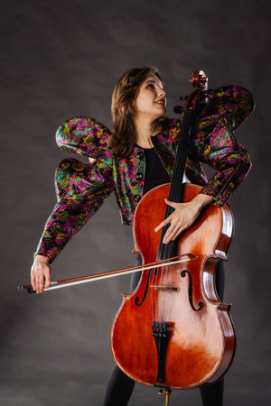 A captivating portrait of cellist Anna Skladannaya against a dark background. She is elegantly dressed in a floral patterned jacket and black pants, holding a cello and bow with grace and focus. The lighting highlights her features and the rich tones of the instrument, creating a dramatic and artistic composition. Perfect for representing music, art, culture, and talent.の写真素材