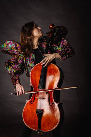 A captivating portrait of cellist Anna Skladannaya against a dark background. She is elegantly dressed in a floral patterned jacket and black pants, holding a cello and bow with grace and focus. The lighting highlights her features and the rich tones of the instrument, creating a dramatic and artistic composition. Perfect for representing music, art, culture, and talent.の写真素材