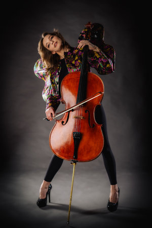 A captivating portrait of cellist Anna Skladannaya against a dark background. She is elegantly dressed in a floral patterned jacket and black pants, holding a cello and bow with grace and focus. The lighting highlights her features and the rich tones of the instrument, creating a dramatic and artistic composition. Perfect for representing music, art, culture, and talent.の写真素材