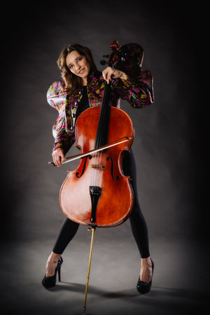 A captivating portrait of cellist Anna Skladannaya against a dark background. She is elegantly dressed in a floral patterned jacket and black pants, holding a cello and bow with grace and focus. The lighting highlights her features and the rich tones of the instrument, creating a dramatic and artistic composition. Perfect for representing music, art, culture, and talent.の写真素材