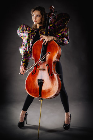 A captivating portrait of a cellist against a dark background. She is elegantly dressed in a floral patterned jacket and black pants, holding a cello and bow with grace and focus. The lighting highlights her features and the rich tones of the instrument, creating a dramatic and artistic composition. Perfect for representing music, art, culture, and talent.の写真素材