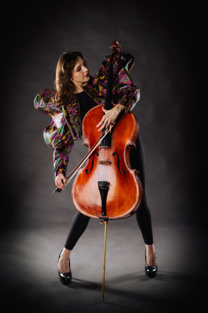 A captivating portrait of a cellist against a dark background. She is elegantly dressed in a floral patterned jacket and black pants, holding a cello and bow with grace and focus. The lighting highlights her features and the rich tones of the instrument, creating a dramatic and artistic composition. Perfect for representing music, art, culture, and talent.の写真素材