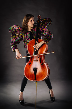 A captivating portrait of cellist Anna Skladannaya against a dark background. She is elegantly dressed in a floral patterned jacket and black pants, holding a cello and bow with grace and focus. The lighting highlights her features and the rich tones of the instrument, creating a dramatic and artistic composition. Perfect for representing music, art, culture, and talent.の写真素材