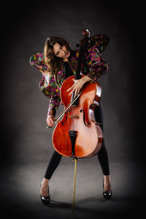 A captivating portrait of cellist Anna Skladannaya against a dark background. She is elegantly dressed in a floral patterned jacket and black pants, holding a cello and bow with grace and focus. The lighting highlights her features and the rich tones of the instrument, creating a dramatic and artistic composition. Perfect for representing music, art, culture, and talent.の写真素材