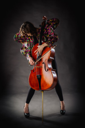 A captivating portrait of a cellist against a dark background. She is elegantly dressed in a floral patterned jacket and black pants, holding a cello and bow with grace and focus. The lighting highlights her features and the rich tones of the instrument, creating a dramatic and artistic composition. Perfect for representing music, art, culture, and talent.の写真素材