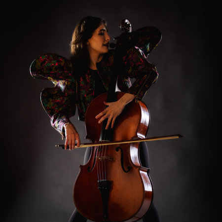 A captivating portrait of cellist Anna Skladannaya against a dark background. She is elegantly dressed in a floral patterned jacket and black pants, holding a cello and bow with grace and focus. The lighting highlights her features and the rich tones of the instrument, creating a dramatic and artistic composition. Perfect for representing music, art, culture, and talent.の写真素材