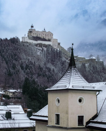 A majestic view of Hohenwerfen Castle perched atop a snow-covered hill. The castle, partially obscured by a light mist, stands as a testament to history and grandeur. The surrounding landscape is blanketed in fresh snow, with bare trees adding to the wintery scene. A truly breathtaking sight.の写真素材