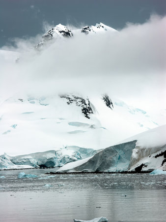 A serene landscape featuring snow-covered mountains partially obscured by clouds, with icebergs floating in a calm body of water.の写真素材