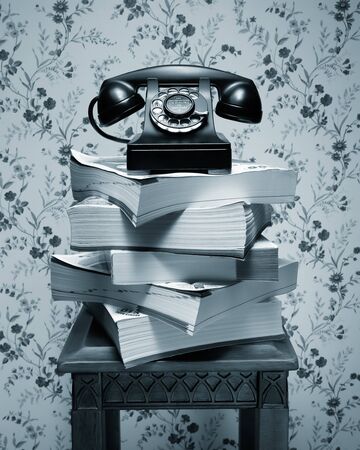 Vintage antique black rotary dial telephone standing on stack of phone books and wooden tableの写真素材
