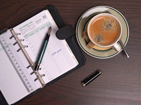 The photo shows a diary, a pen and a cup of coffee on a tableの写真素材