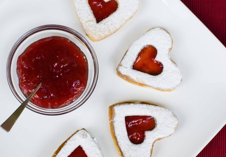 Heart shape strawberry jam cookies placed on a white plate with jam and red background