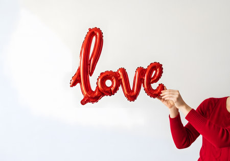 A person holds a large red cursive love foil balloon against a bright white background, ideal for celebrations and greetings.の素材