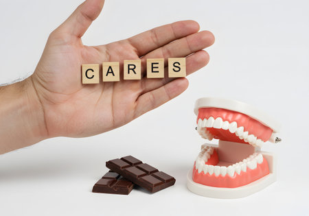 A hand holds wooden tiles spelling caries (dental decay) above a jaw model and a pile of chocolate. The visual represents the negative effect of sugar and sweets on dental health and oral hygiene.の素材