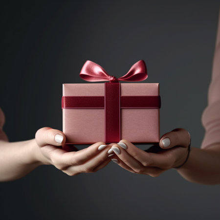 Two hands holding a gift box with red bow on dark background.の素材