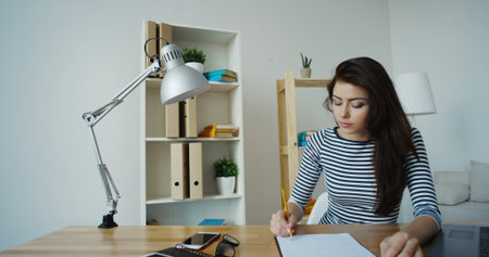 Confident brunette businesswoman in striped blouse and glasses taking note in a notepad at office