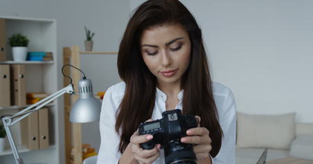 Smiling brunette woman wearing white shirt holding camera and looking at you, office interior at background.の写真素材