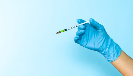 Hand in blue glove holding a plastic syringe with liquid for injection on light blue background....
