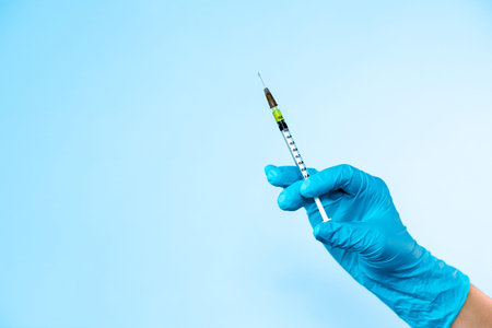 Hand in blue glove holding a plastic syringe with liquid for injection on light blue background....