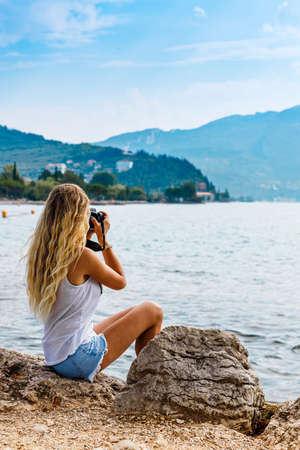 The girl sitting near water with photo camera. Blonde takes pictures of the mountains. Back view, vacation conceptの写真素材