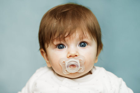 Close up portrait of five-month-old baby girl with a pacifier in her mouth looking at camera in blue...
