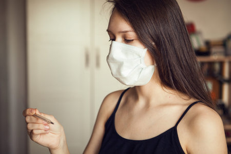 Young girl with ear loop face mask looking at medical thermometer in hand, close up