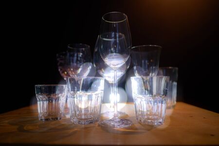 Close ups of wine-, long drink- and spirits-glasses on a wooden shelve in a bar, in front of a dark background, illuminated by colorful spot lights, creating blurry light effects with beautiful  bokehの写真素材