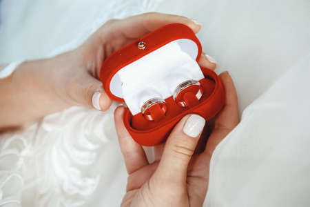 The bride holds two wedding rings in a red velvet box on the background of a white wedding dressの写真素材