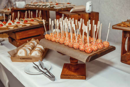 candies in multi-colored sprinkles on sticks against the background of trays with sandwiches and buns in glaze on a table with a white tableclothの写真素材