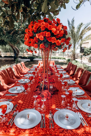 red table wedding arrangement of roses, carnations on the background of a table with a red tablecloth, red chairs and a tree in summerの写真素材