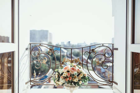 a flowerpot with flower arrangements of pink and white roses, white hydrangea and sprigs of greenery for bouquets stands on a balcony with doors wide open against the background of the cityの写真素材