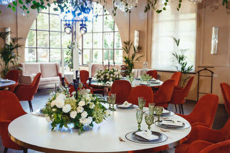 restaurant hall with white tables and red exquisite chairs, decorated with floral arrangements on the tables, homemade exotic plants, designer chandeliers and greeneryの写真素材