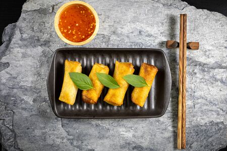 4 fried spring rolls in a black ceramic plate and wooden chopsticks placed on a stone plate. Top view.の写真素材