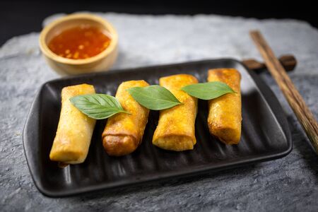 4 fried spring rolls in a black ceramic plate and wooden chopsticks placed on a stone plate. Close up shot.の写真素材