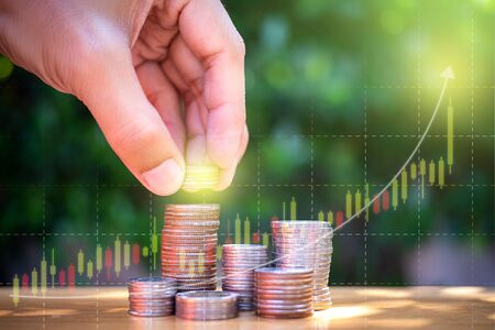 Coin stack with man left hand want to put coin and investing graph background. Saving for the future. Close up shot.の写真素材