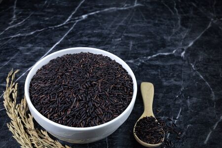 Thai black rice, rice berry  in white ceramic bowl and wooden spoon on dark marble table.の写真素材