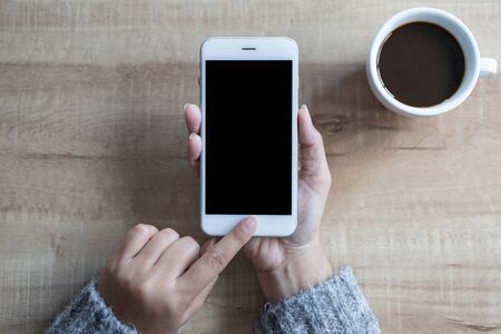 Hand using white smart phone black screen on top view, with white coffee cup on wooden table.の写真素材