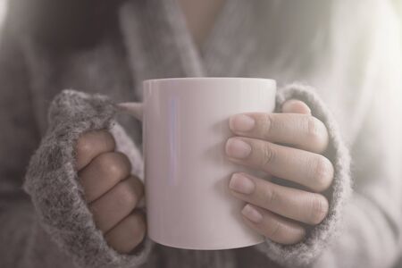 Close up of a woman hand holding a coffee mug in the morning soft sunlight. Beautiful girl in grey sweater.の写真素材