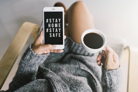 Woman's hands holding black screen mobile phone with text hashtag #STAY HOME and #STAY SAFE and coffee cup in her hand. Concept stay home stop virus.の写真素材