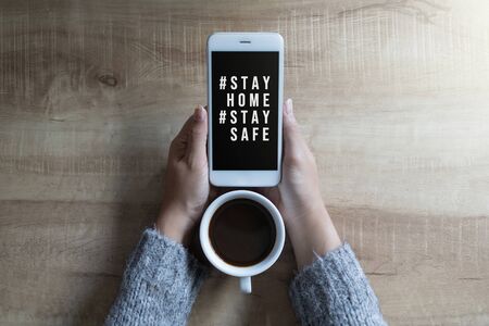 Woman's hands holding black screen mobile phone with text hashtag #STAY HOME and #STAY SAFE and coffee cup in her hand. Concept stay home stop virus.の写真素材