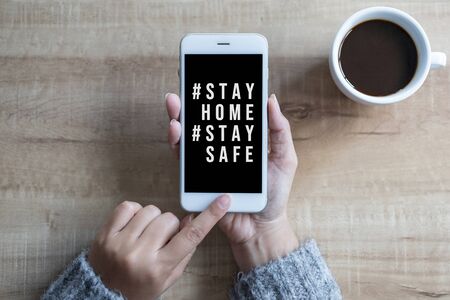 Woman's hands holding black screen mobile phone with text hashtag #STAY HOME and #STAY SAFE and coffee cup in her hand. Concept stay home stop virus.の写真素材